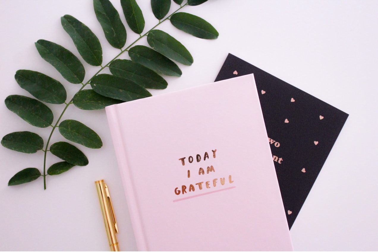 a soft pink notebook with the words ‘today I am grateful’ sitting on a table with a gold pen and leaf