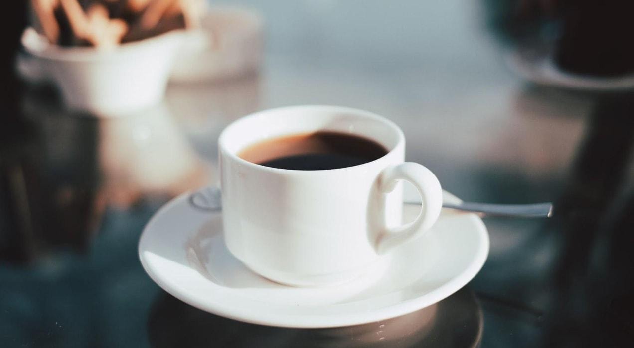 A reflective glass table with a short white coffee cup filled with black coffee, sitting on a white saucer