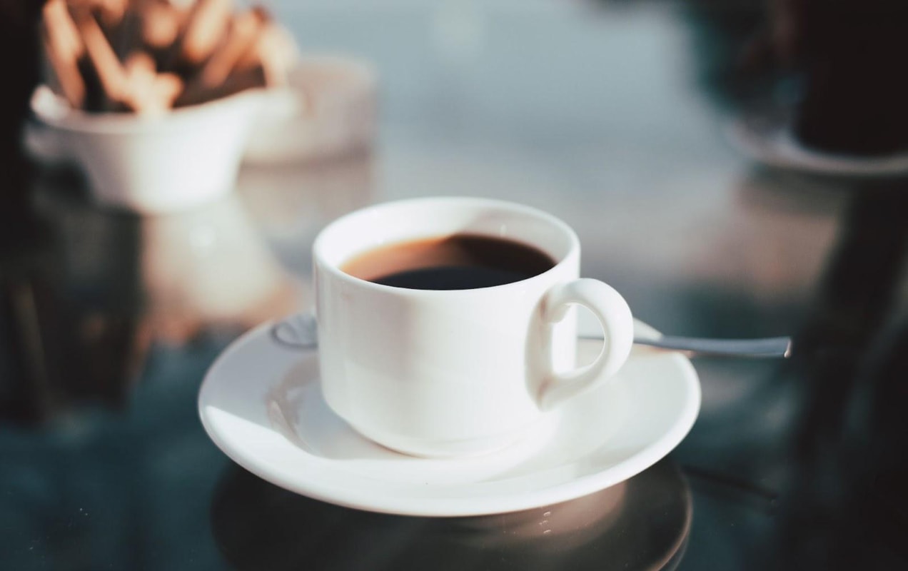 A reflective glass table with a short white coffee cup filled with black coffee, sitting on a white saucer