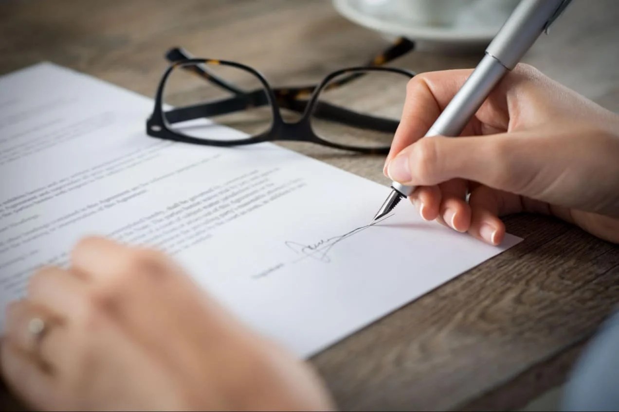 A person signing a contract with their glasses in the background.