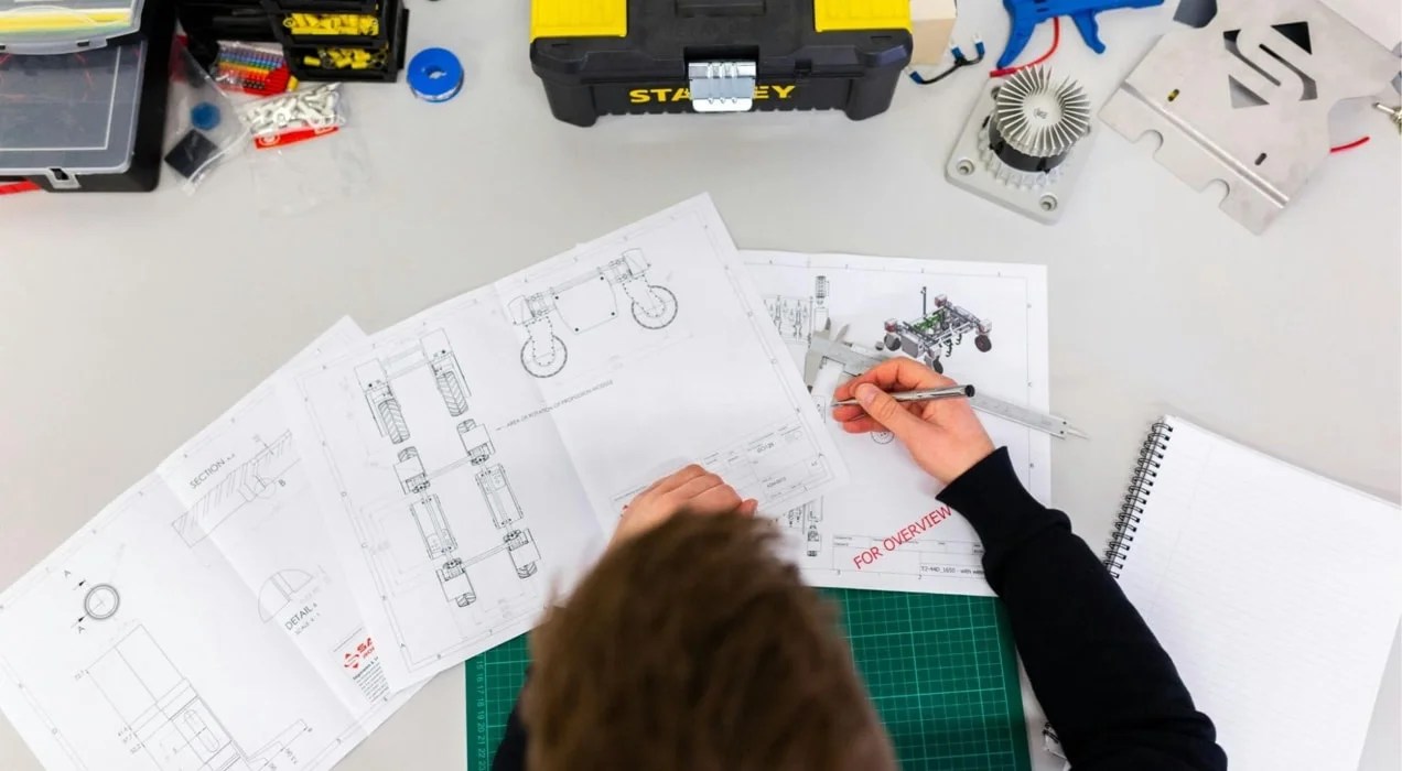 someone sketching equipment parts at a desk with a toolbox